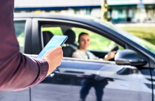 Person holding a smartphone to book a rideshare while the driver waits in the car.