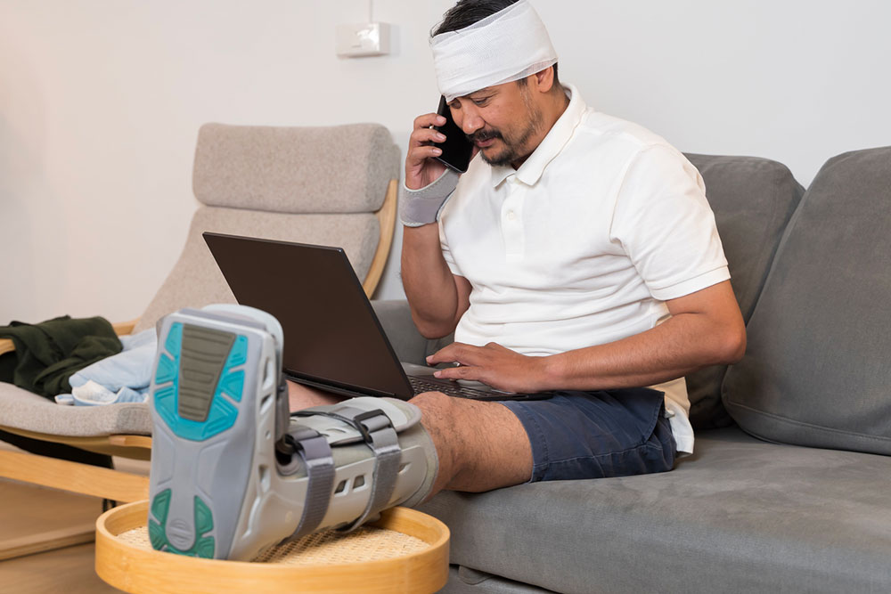 Injured man with a leg brace and head bandage using a laptop and talking on the phone during home recovery.