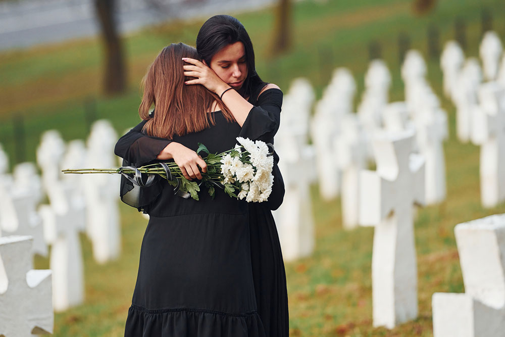 Two women in black embrace and cry at a cemetery during a funeral visit.