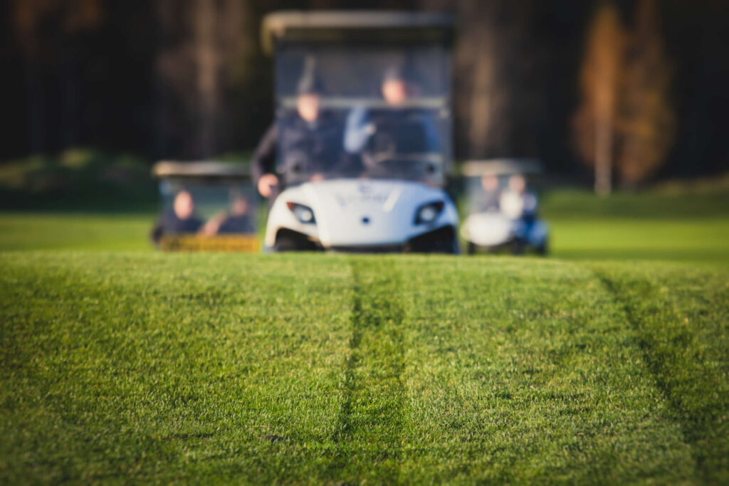 White golf cart driving across a grassy fairway with tire tracks in the foreground.
