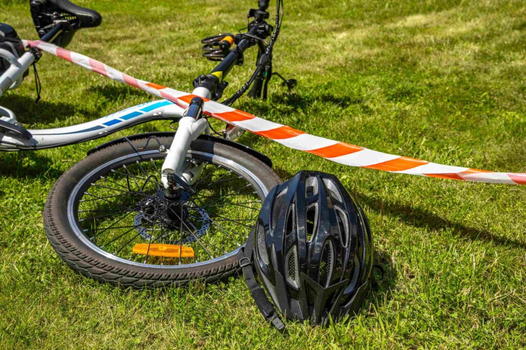 A black bicycle helmet lies on the green grass next to a fallen bicycle wheel wrapped in orange and white caution tape.