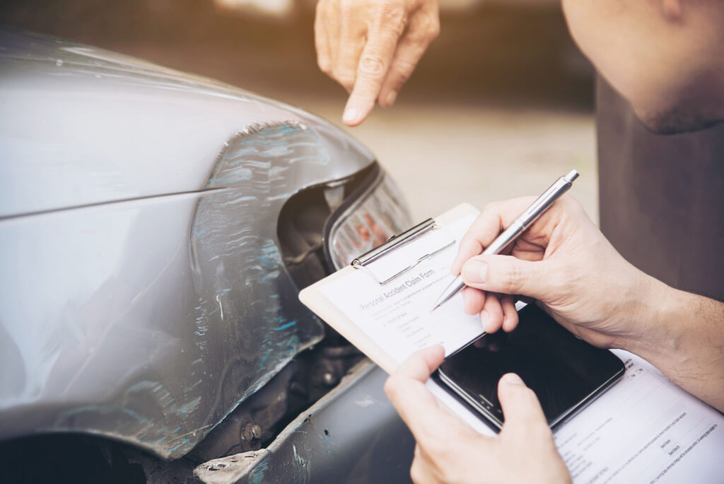 Close-up of a person filling out a personal accident claim form on a clipboard next to a dented and scratched grey vehicle bumper while another hand points at the damage.