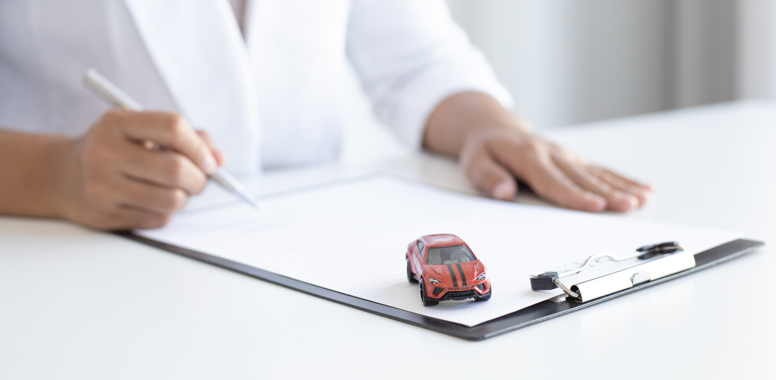 A person holding a pen over a clipboard with paperwork and a small red toy model car sitting on the document.