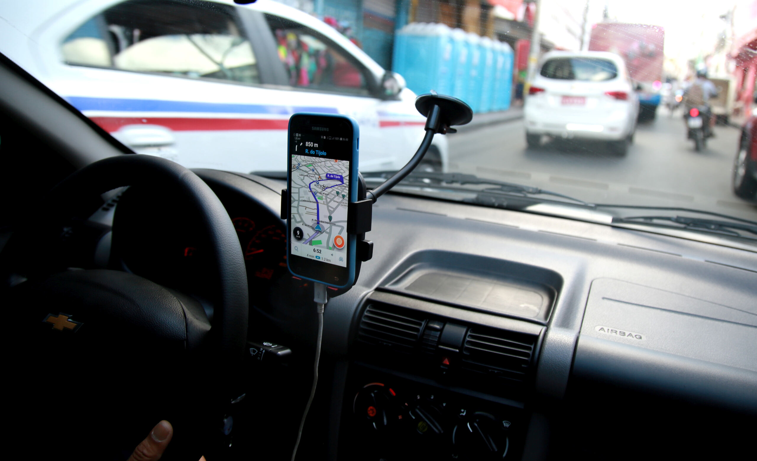 View from inside a vehicle dashboard showing a smartphone mounted on the windshield displaying a GPS navigation map application during a drive on a city street.