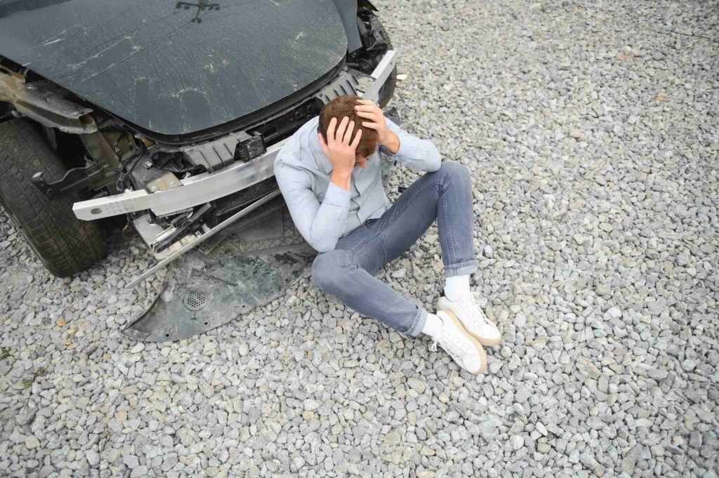 A man sitting on the ground holding his head next to a crashed vehicle highlights injuries leading to Safety Harbor Concussion & Mild TBI Claims.
